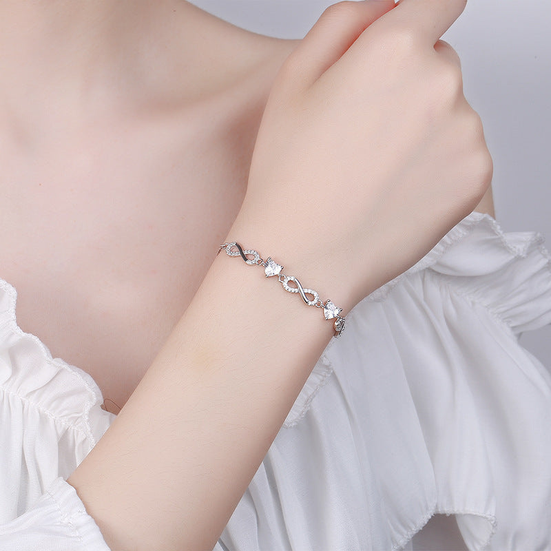 Hand wearing a silver bracelet with heart-shaped charms on a light Silver bracelet with infinity symbols Aru Collectionbackground